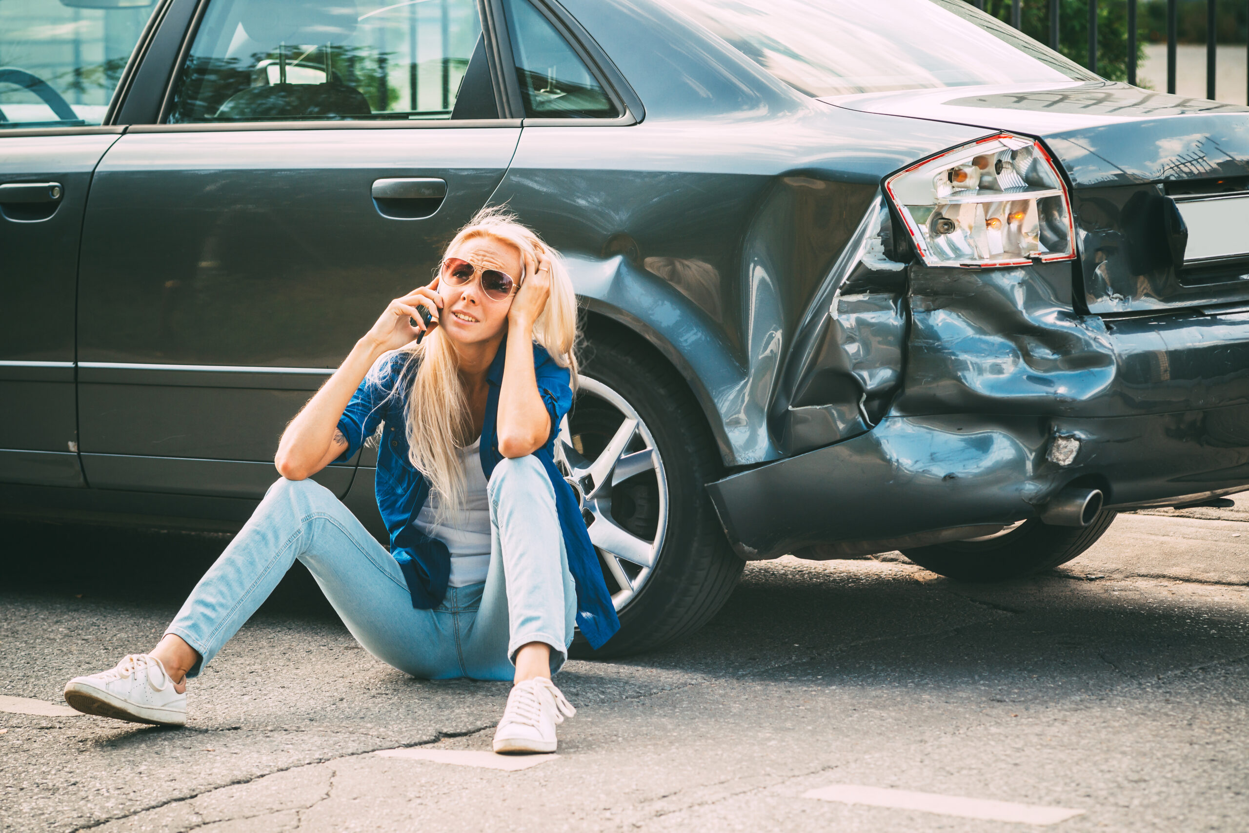 A girl sits on the road, near the broken car, and calls her car insurance company for help.