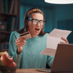 A shocked woman holding a credit card, staring at a paper bill in shock as she discovers excessive card surcharges