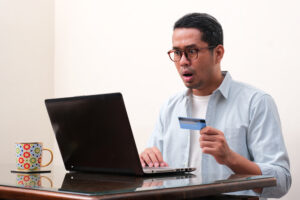 A man who has been scammed looks at his laptop with a shocked expression while holding his credit card