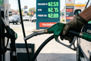 Close-up of a hand holding a fuel nozzle while refuelling, with two nearby petrol stations in the blurred background showing a clear fuel price gap between them