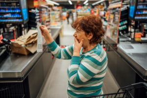 Elderly woman looking shocked while reading a supermarket receipt, reacting to unexpected grocery prices or discount changes.