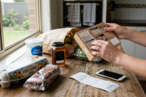 Person checking food labels and ingredients on everyday groceries spread across a kitchen bench, with a receipt and smartphone nearby.